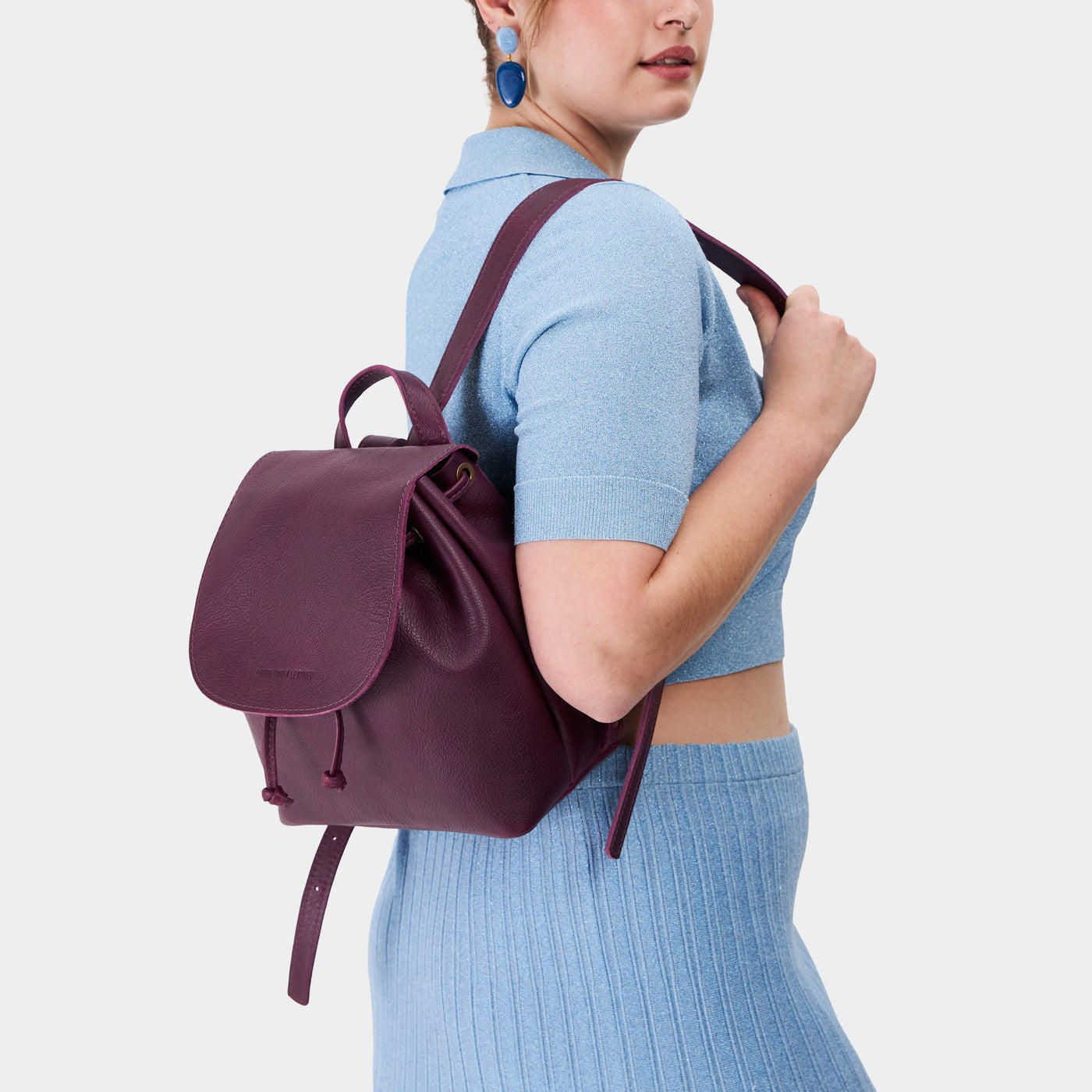 Plum | Pebbled purple leather drawstring backpack being worn by a person wearing a blue sweater.