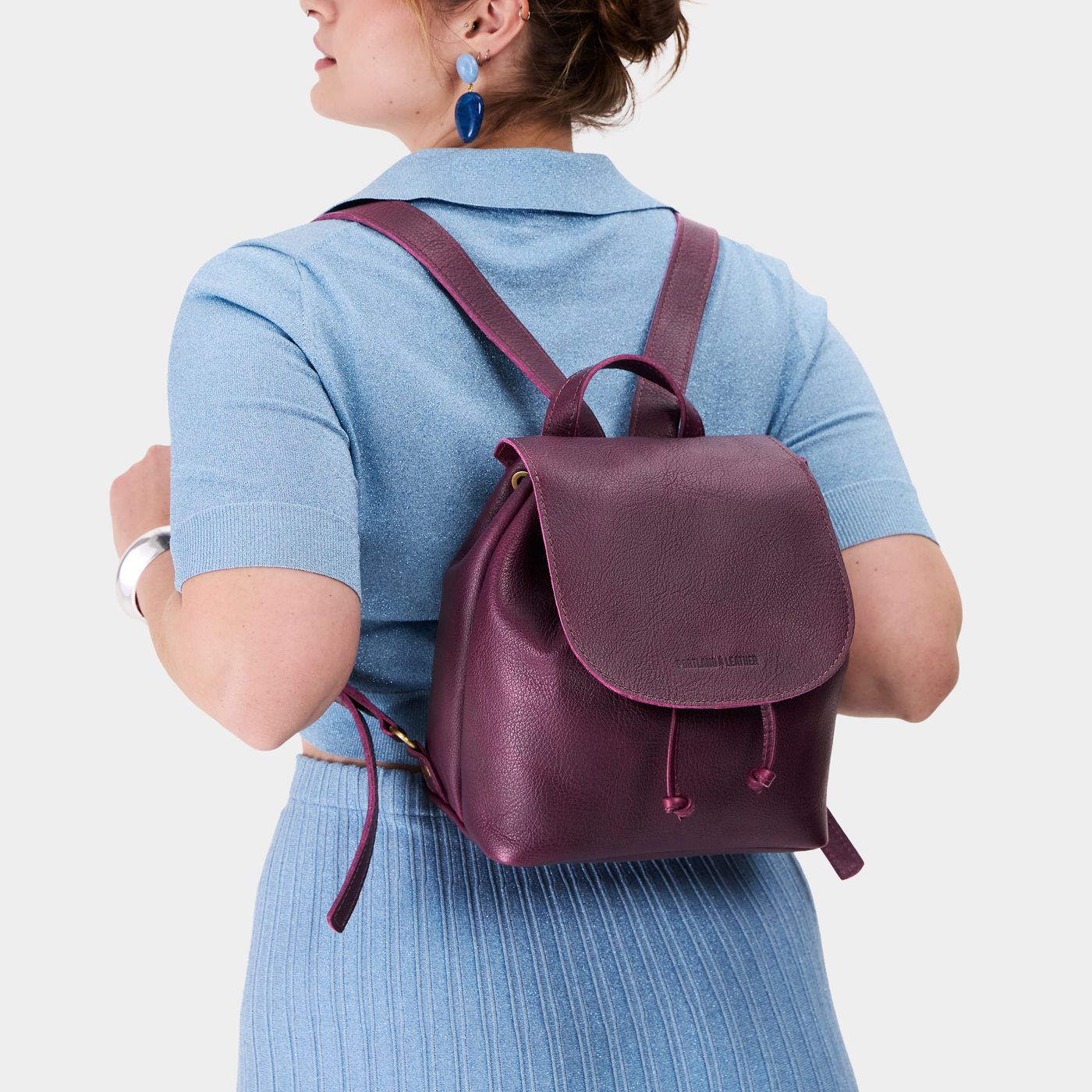 Plum | Pebbled purple leather drawstring backpack being worn by a person wearing a blue sweater.