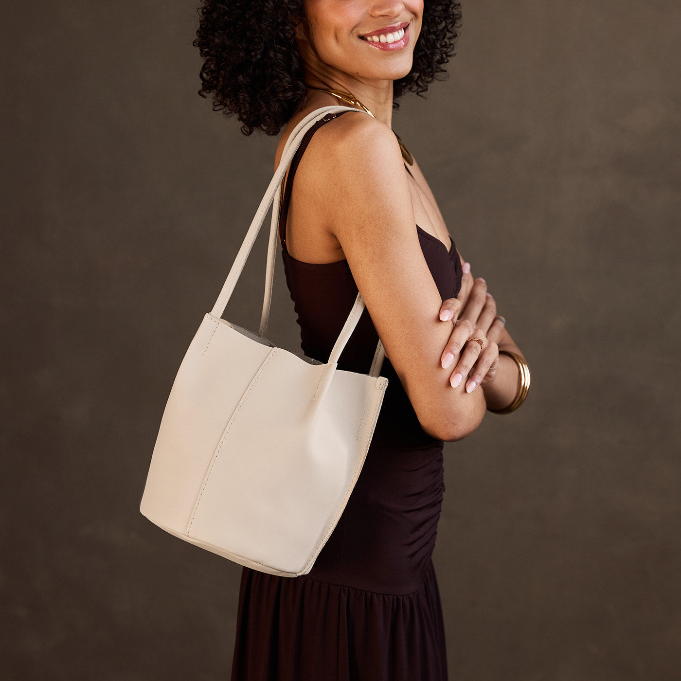 Bone*Small | Model wearing sleeveless dark brown dress carrying petite bucket shaped tote bag with matching leather handles against dark grey background