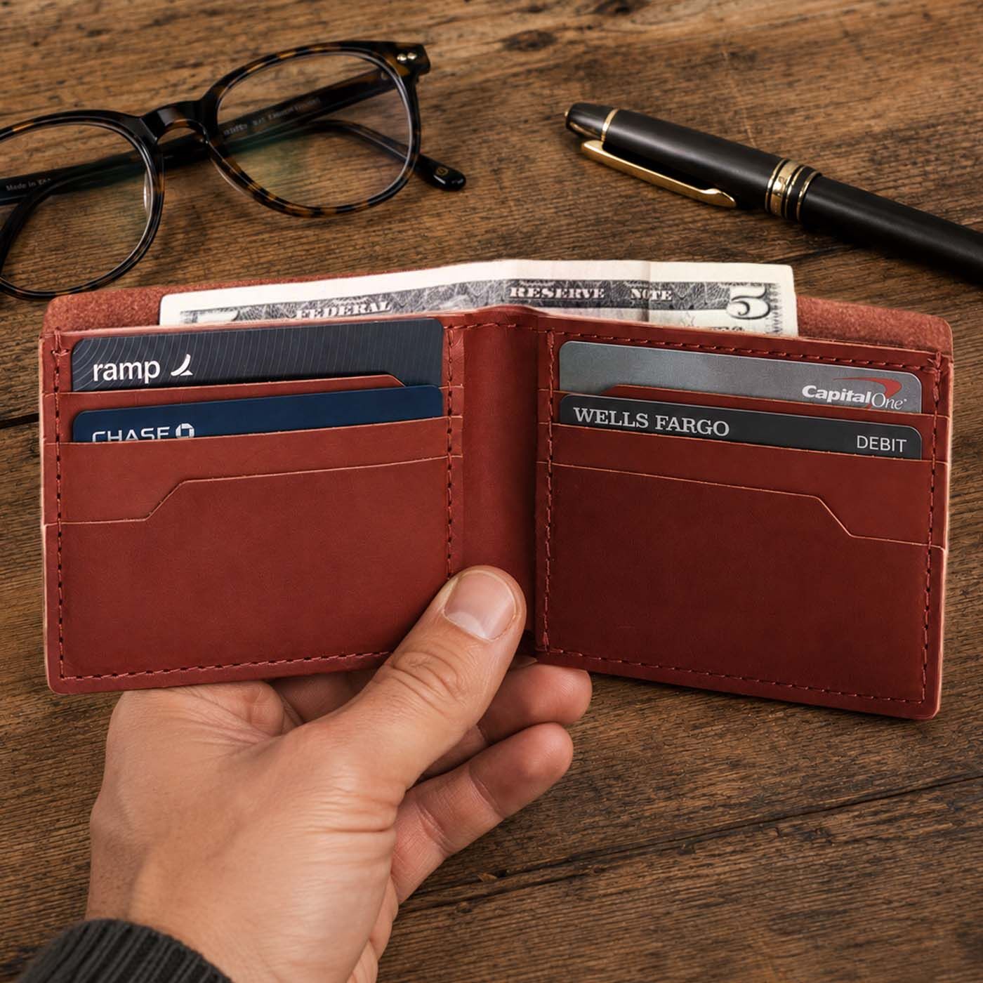 Marrone | A hand holds an open leather bifold wallet showing card slots and cash on a wooden tabletop in the background