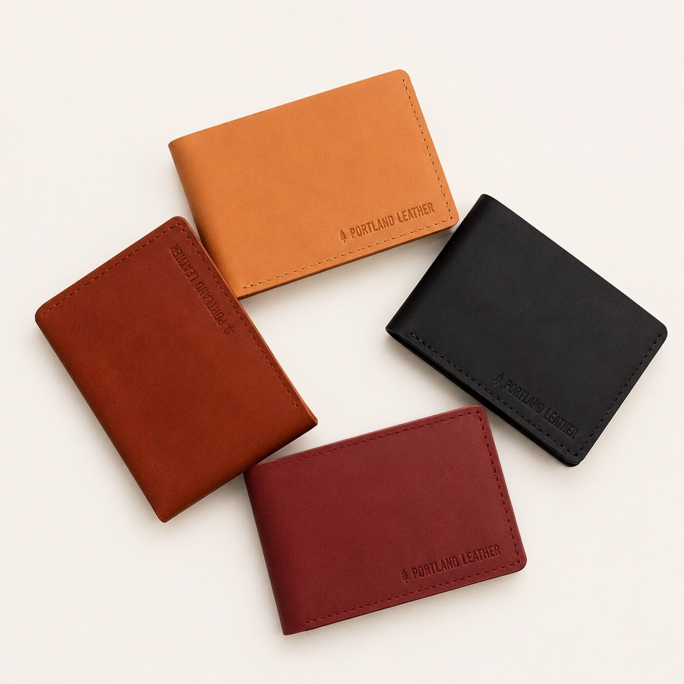 Marrone | Four leather bifold wallets in Naturale, Toscano, Nero, and Marrone, arranged on a light cream background. Each wallet features a debossed logo reading 'PORTLAND LEATHER' in the bottom right corner.