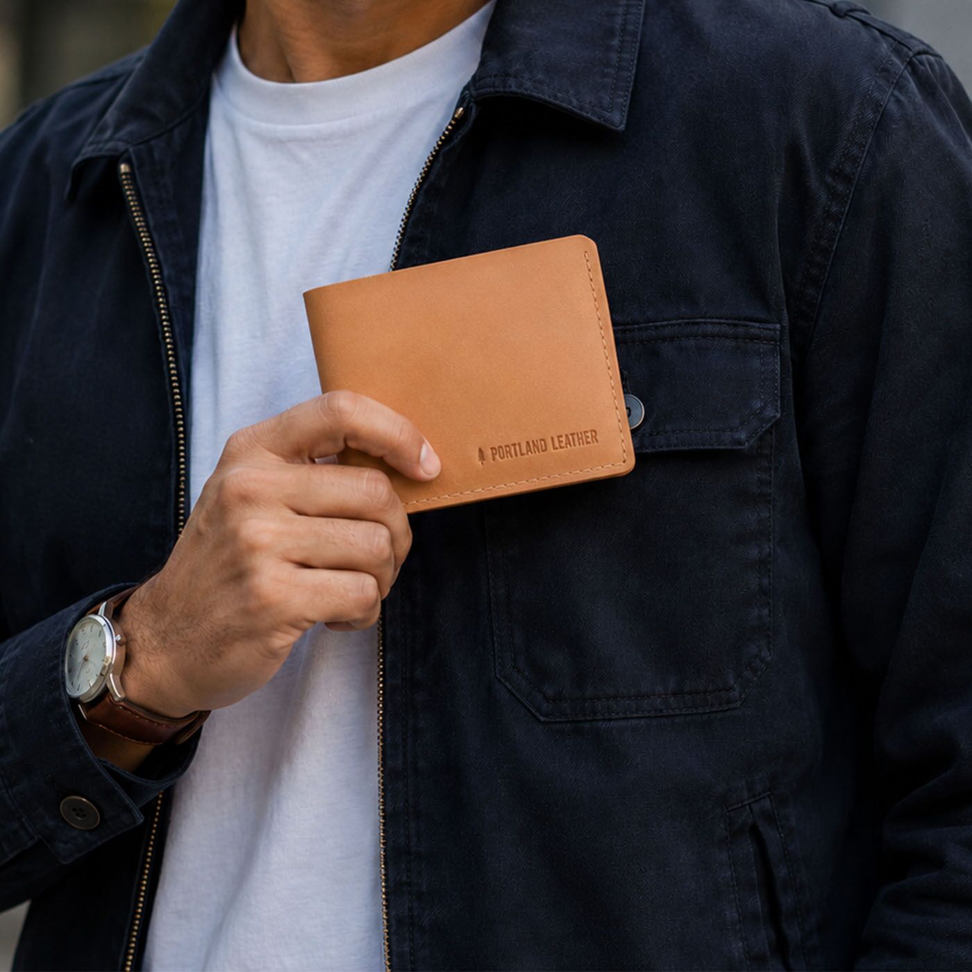 Naturale | A man wearing a dark navy jacket over a white t-shirt holds a Naturale leather bifold wallet with 'PORTLAND LEATHER' embossed on the front.