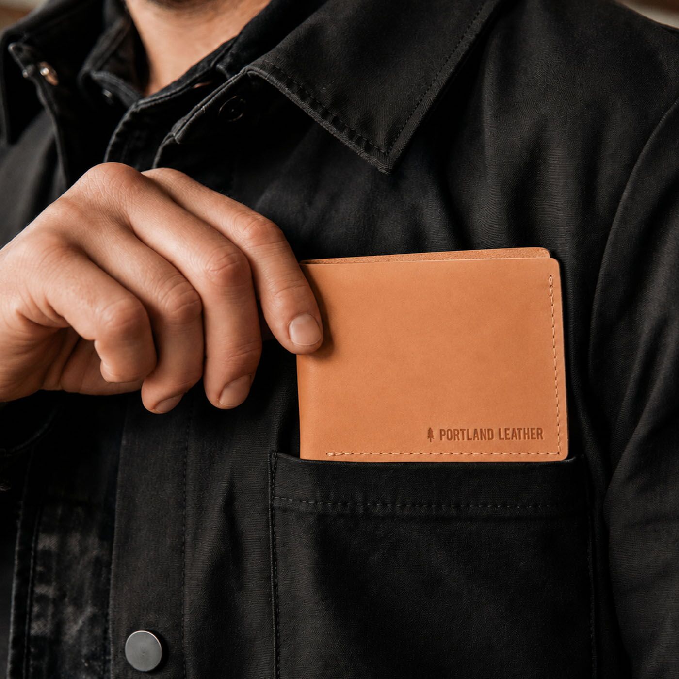 Naturale | A close-up of a person wearing a black overshirt and holding a leather bifold wallet in their hand. The wallet has 'PORTLAND LEATHER' debossed on the bottom right corner.