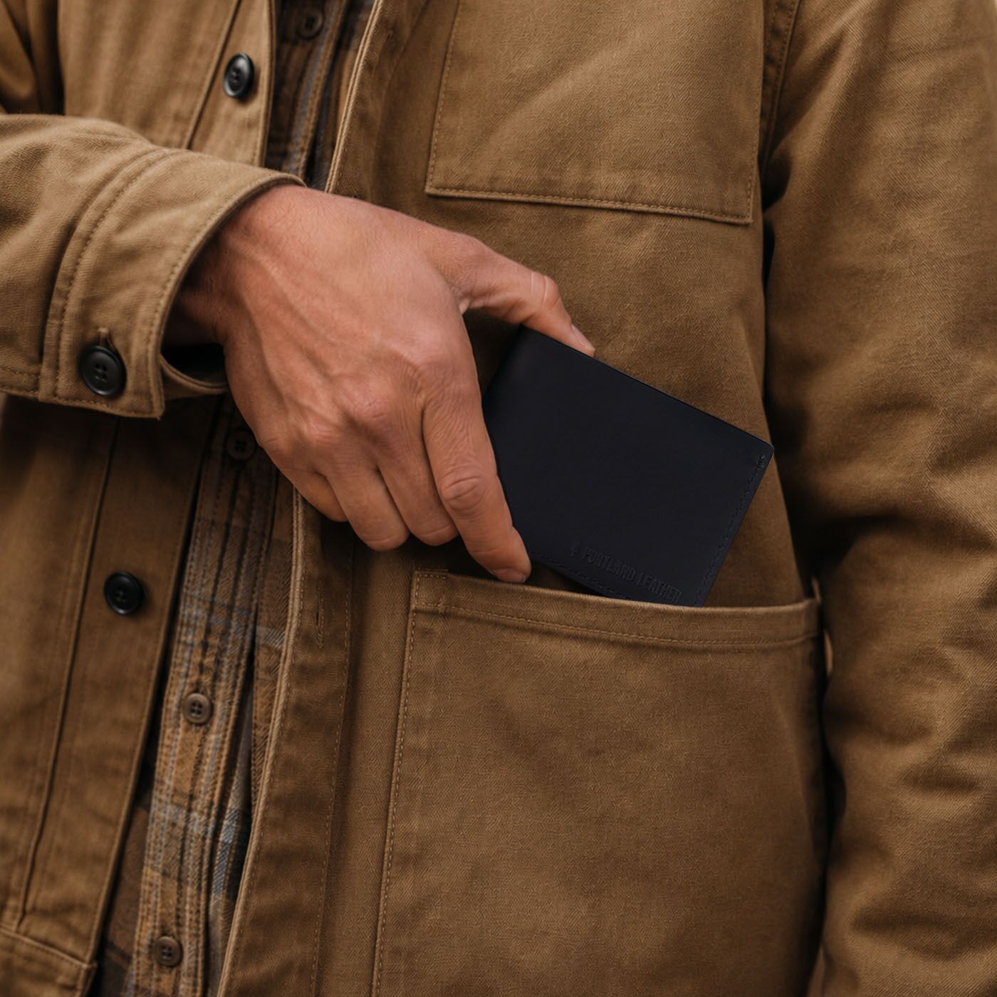 Nero | A close-up of a person wearing a brown waxed canvas utility jacket, showing a hand sliding a Nero bifold wallet into the front utility pocket.