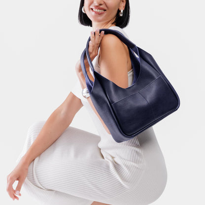 Cobalt*Medium | A model holding a u-shaped, flat-bottomed leather shoulder bag with two outside pockets