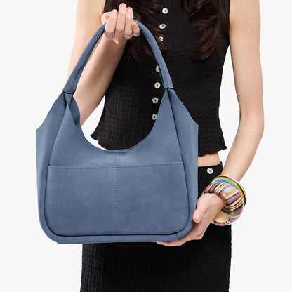 Denim*Medium | A model holding a u-shaped, flat-bottomed leather shoulder bag with two outside pockets