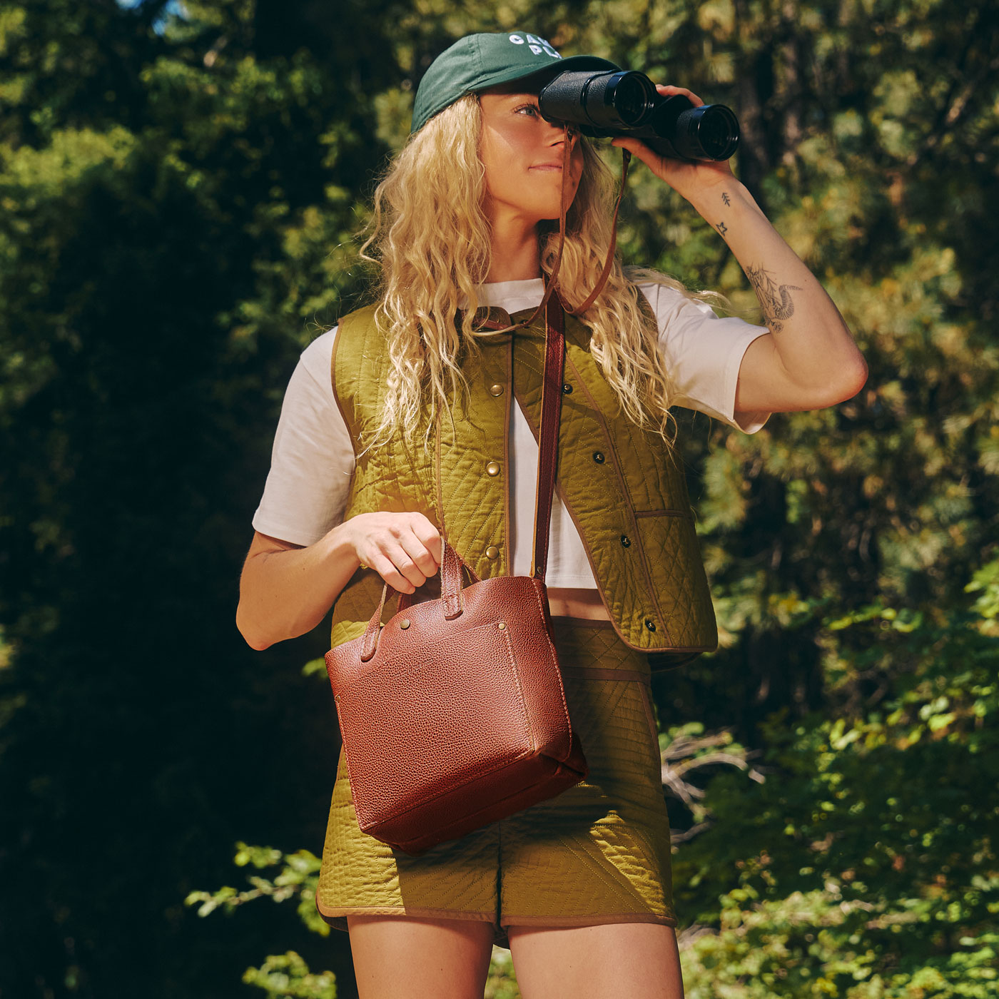 A woman outdoors wearing a green cap, olive green quilted vest and shorts over a white t-shirt, holding a Nutmeg Mini crossbody bag and looking through binoculars.