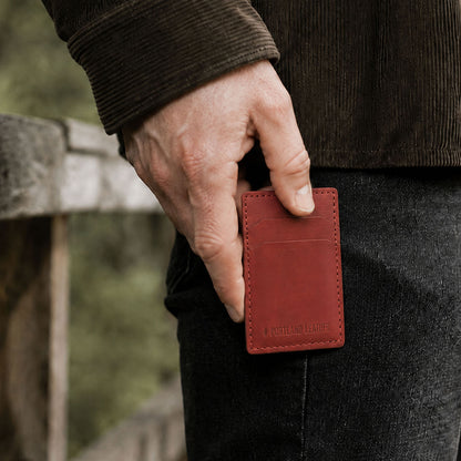 Marrone | A close-up of a model's hand holding a small red leather card holder against their dark jeans, with a blurred outdoor background.