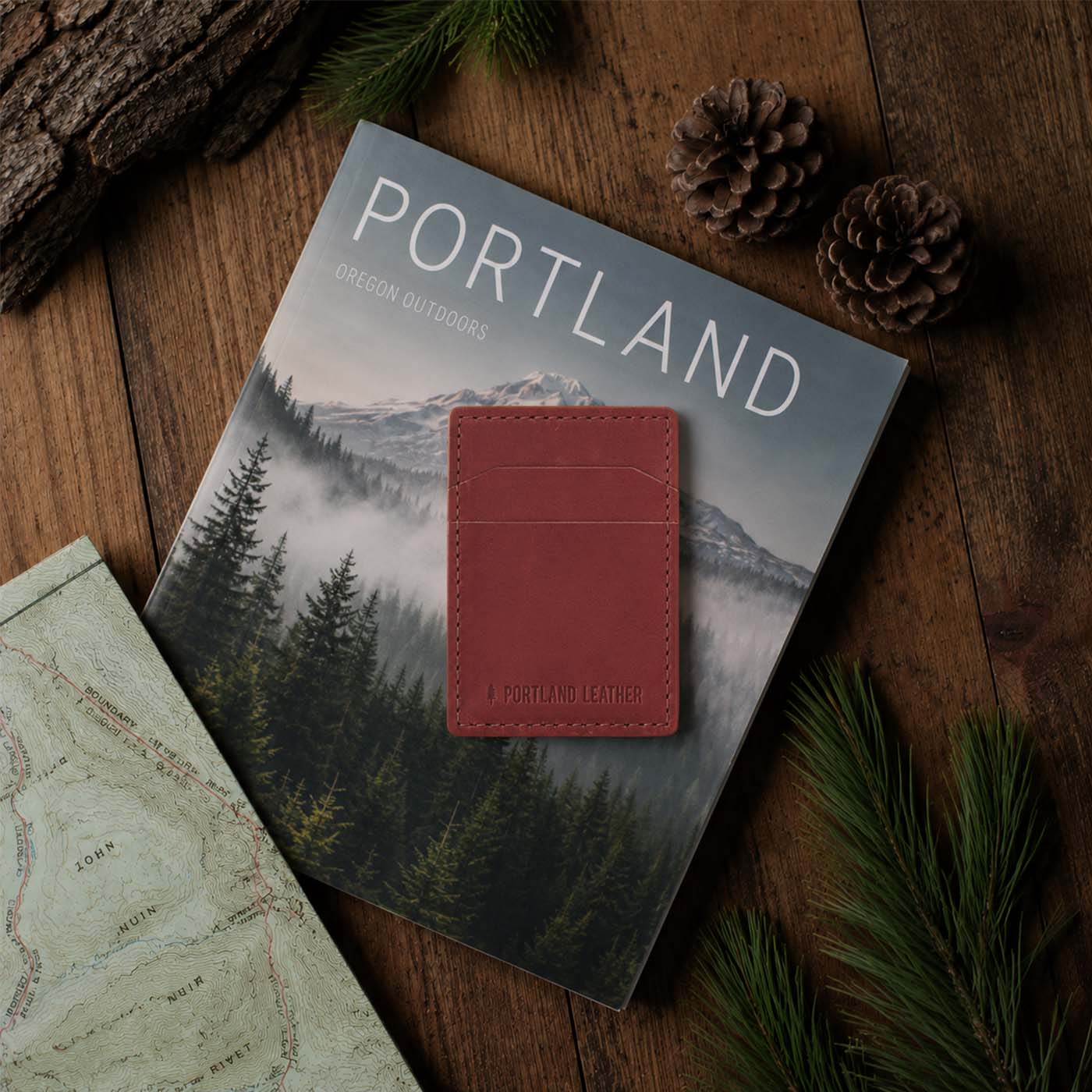 Marrone | A leather card holder wallet rests on top of a Portland Outdoors magazine, styled on a rustic wooden table with tree branch, pinecones, and a map.