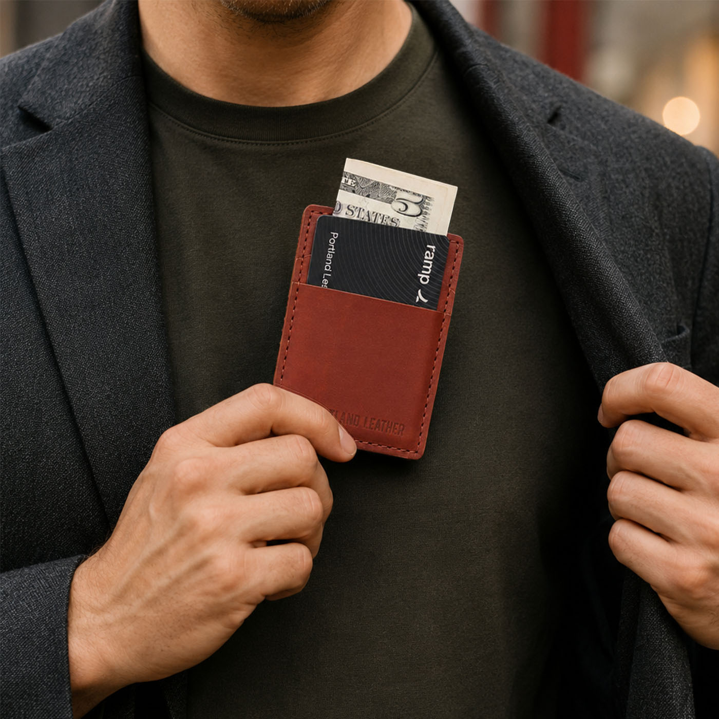 Marrone | A close-up of a model wearing a dark gray blazer over a dark black t-shirt, holding a leather card holder containing a card and cash in front of his chest.
