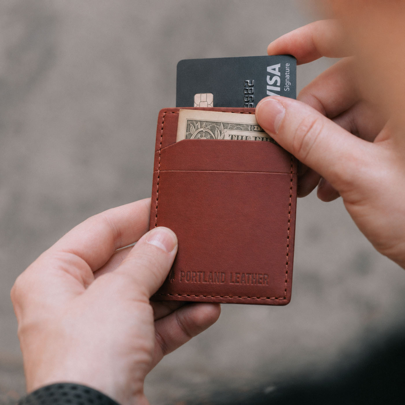 Marrone | A close-up of hands holding a brown leather card holder with a debossed Portland Leather logo, showing card slots containing a black card and a dollar bill, against a blurred gray background.