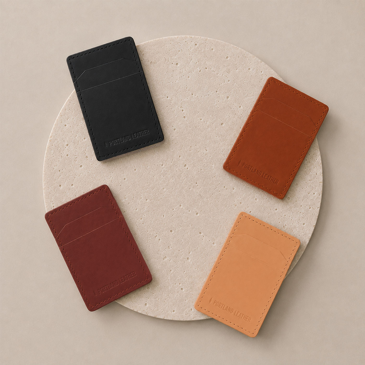 Marrone | Four slim leather card holders are arranged on a round beige textured tray against a neutral beige background. Each holder has a small debossed logo at the bottom.