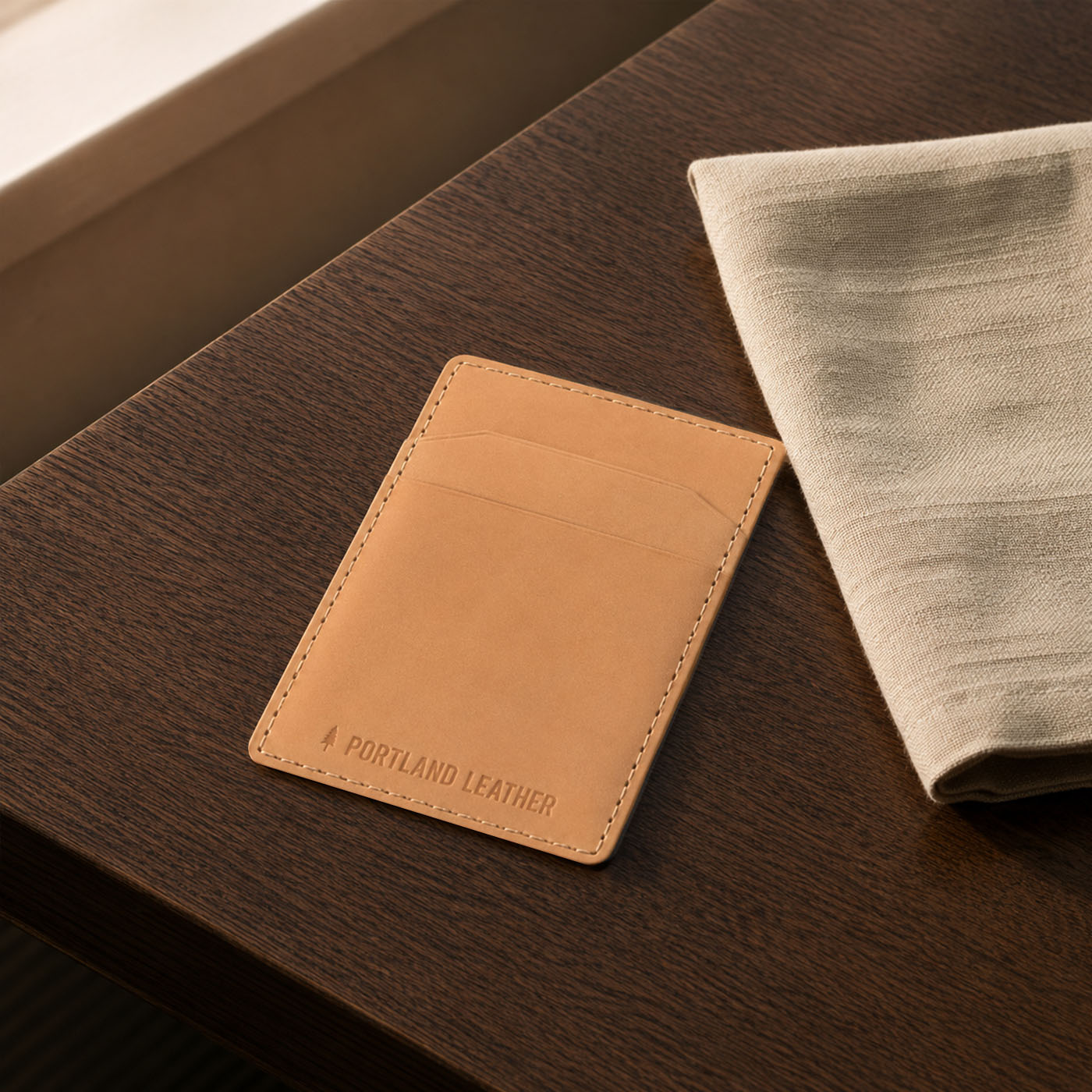 Naturale |A slim tan leather card holder wallet with 'PORTLAND LEATHER' debossed on the front, resting on a dark wood table next to a folded linen cloth.