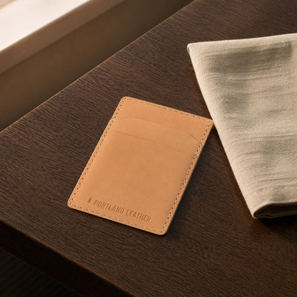 Naturale |A slim tan leather card holder wallet with 'PORTLAND LEATHER' debossed on the front, resting on a dark wood table next to a folded linen cloth.