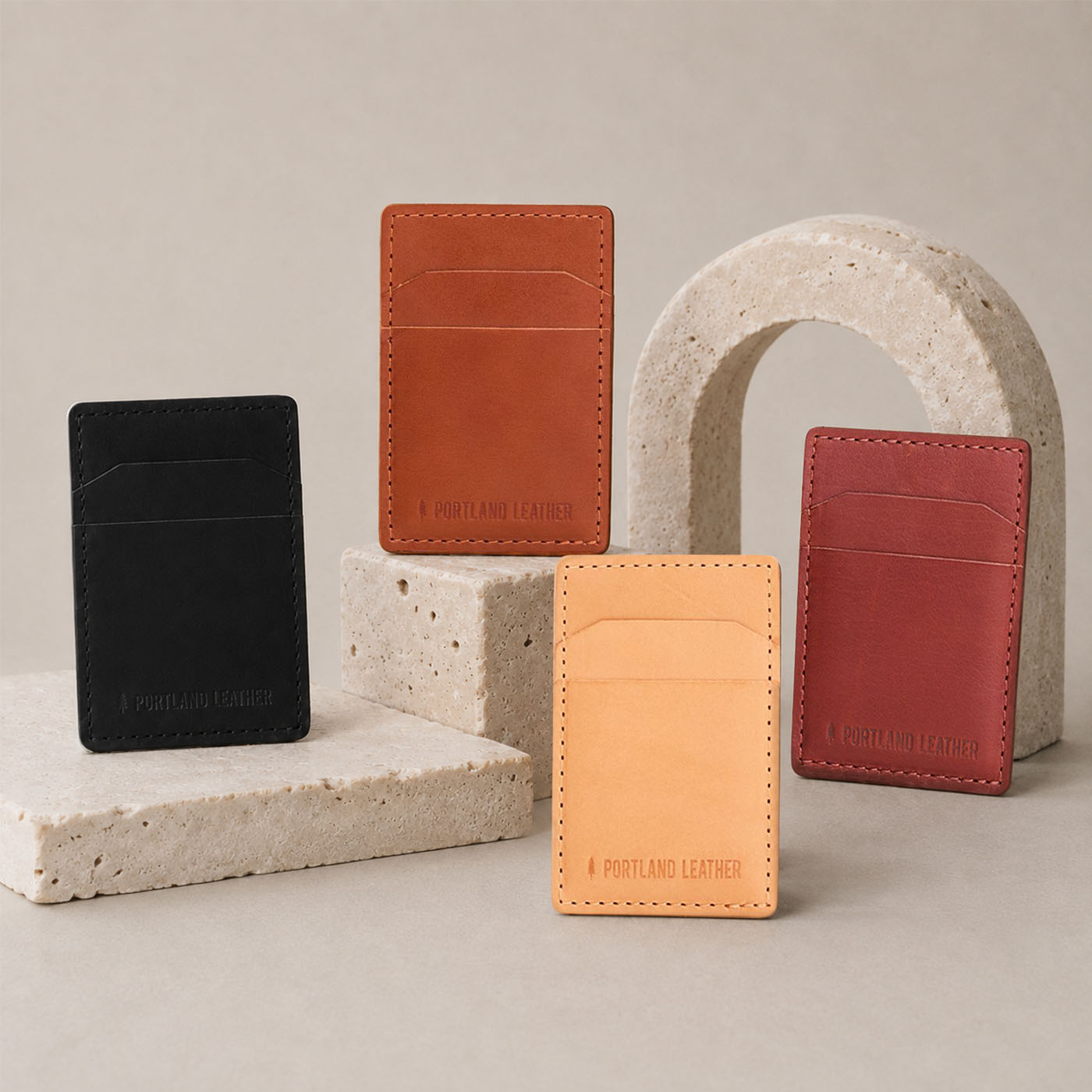Naturale | Four slim leather card holders in black, tan, brown, and rdark brown, arranged on and around light stone blocks and an arch against a warm beige studio background.
