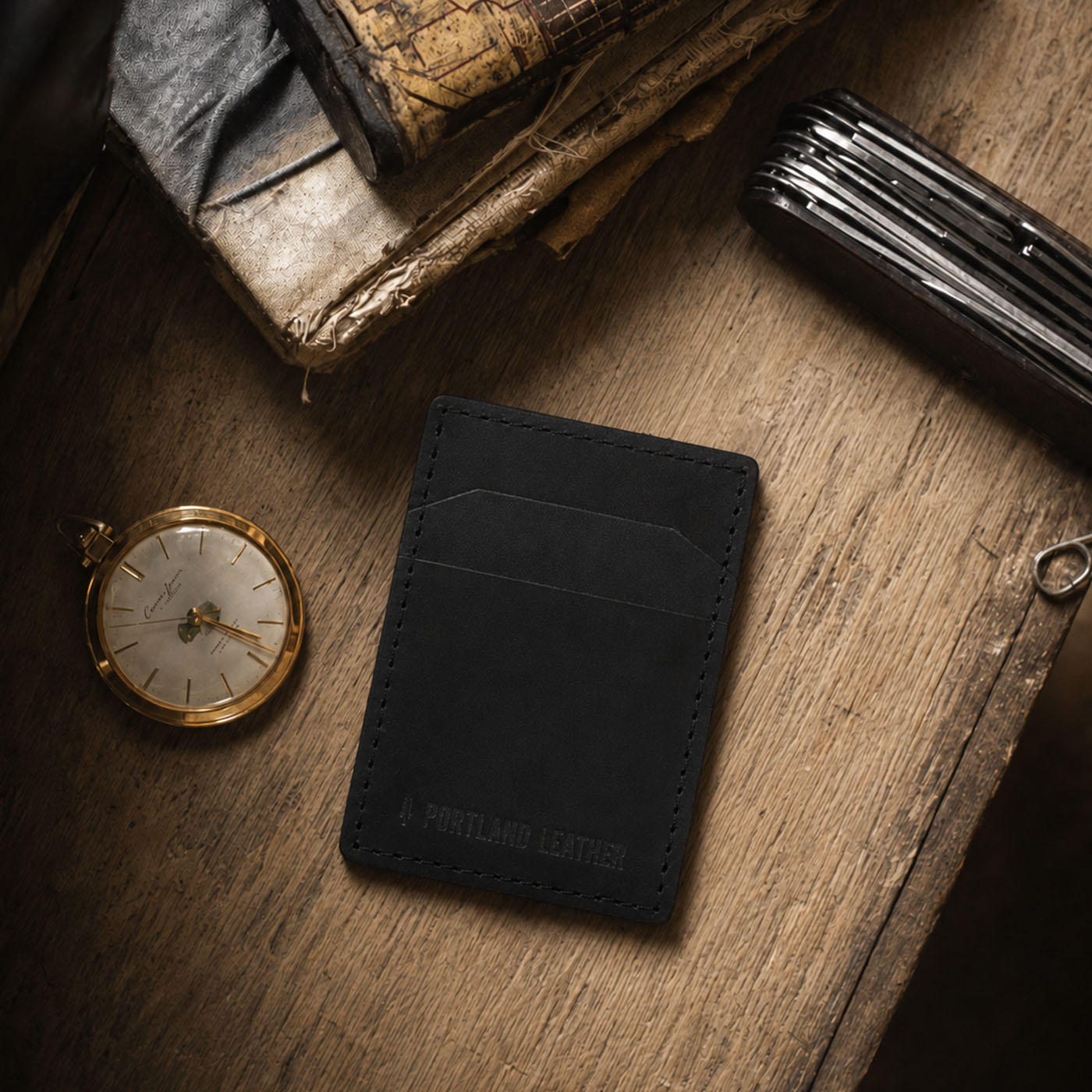 Nero | A black leather card holder styled on a rustic wooden table with vintage books, a pocket watch, and a folding knife.