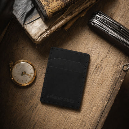 Nero | A black leather card holder styled on a rustic wooden table with vintage books, a pocket watch, and a folding knife.
