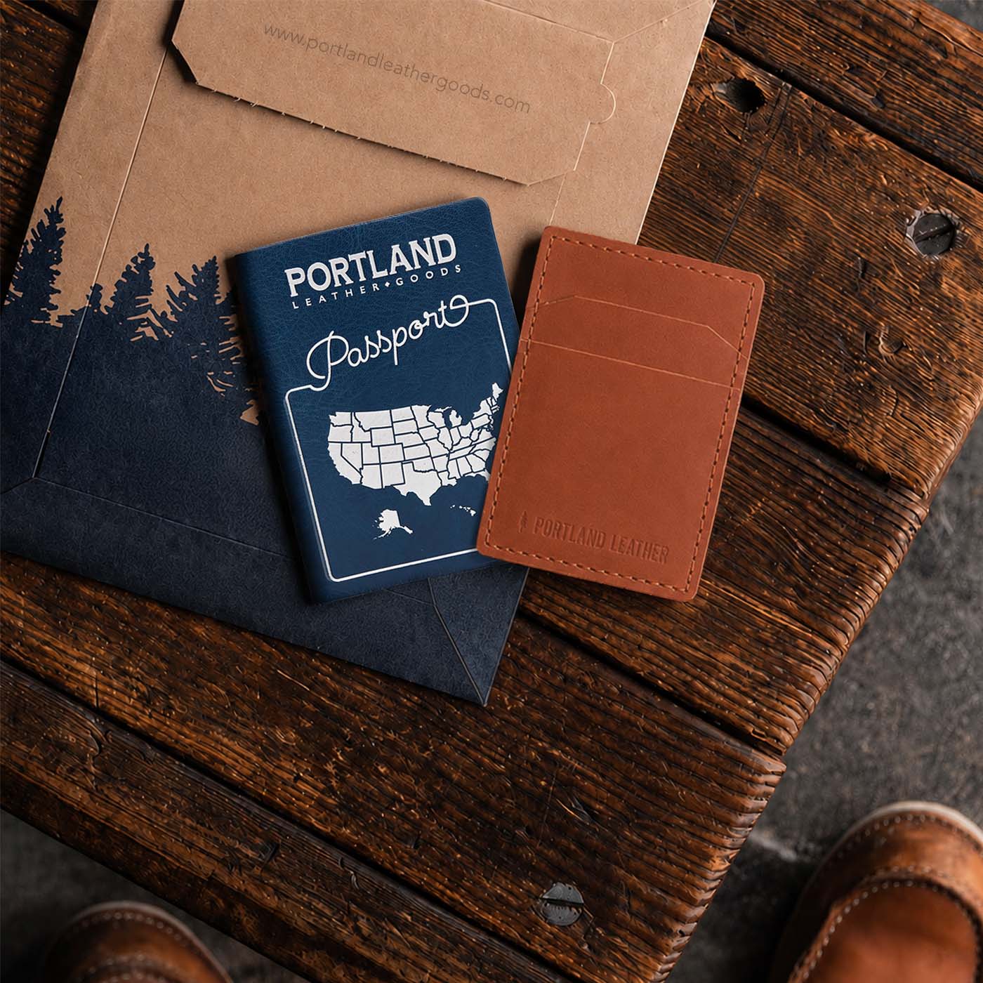 Toscano | A brown leather card holder with a debossed logo, displayed next to a navy blue Portland Leather Goods passport and kraft paper envelope on a rustic wooden table.