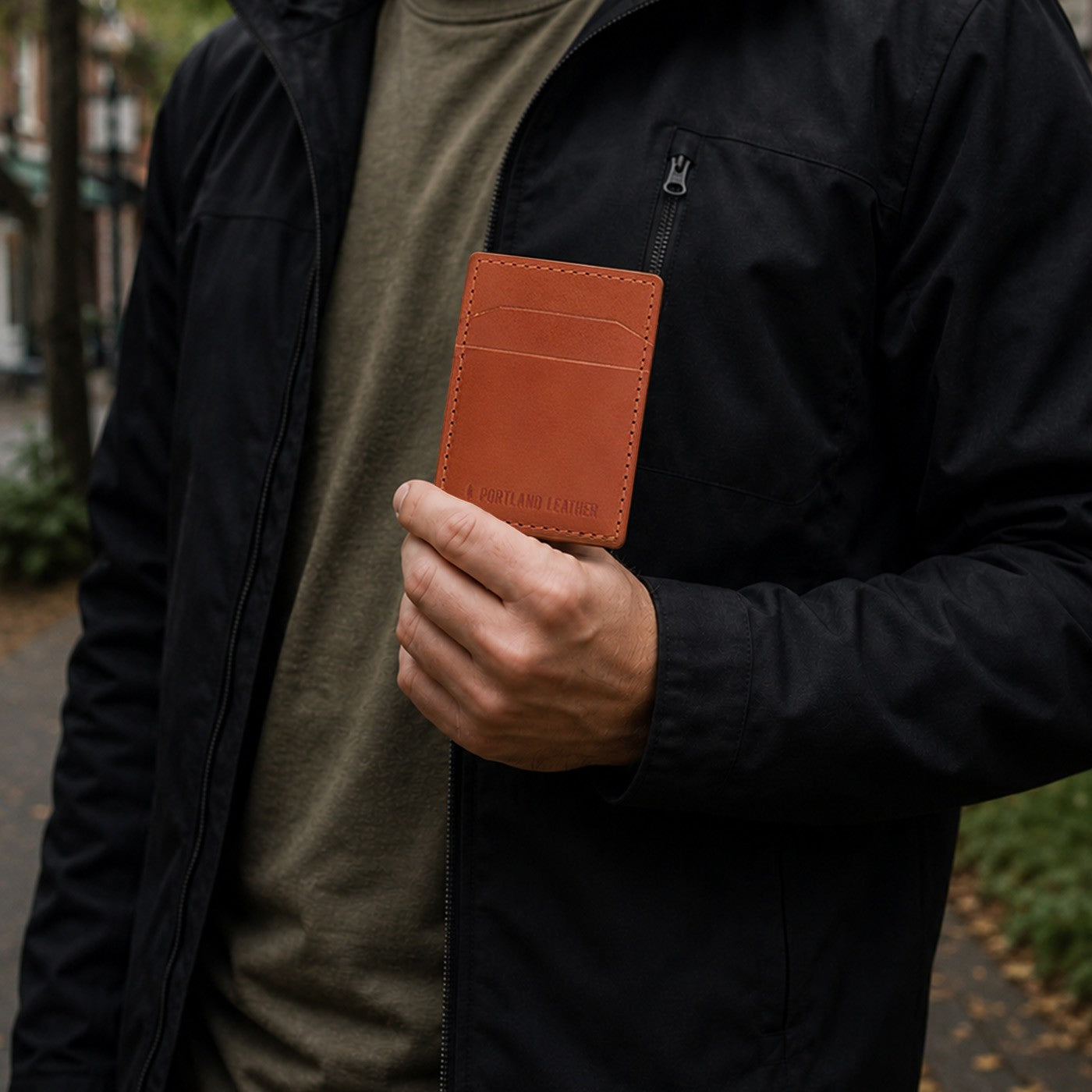 Toscano | A man wearing a black jacket and olive green shirt holds a brown leather card holder in his hand in an outdoor setting.