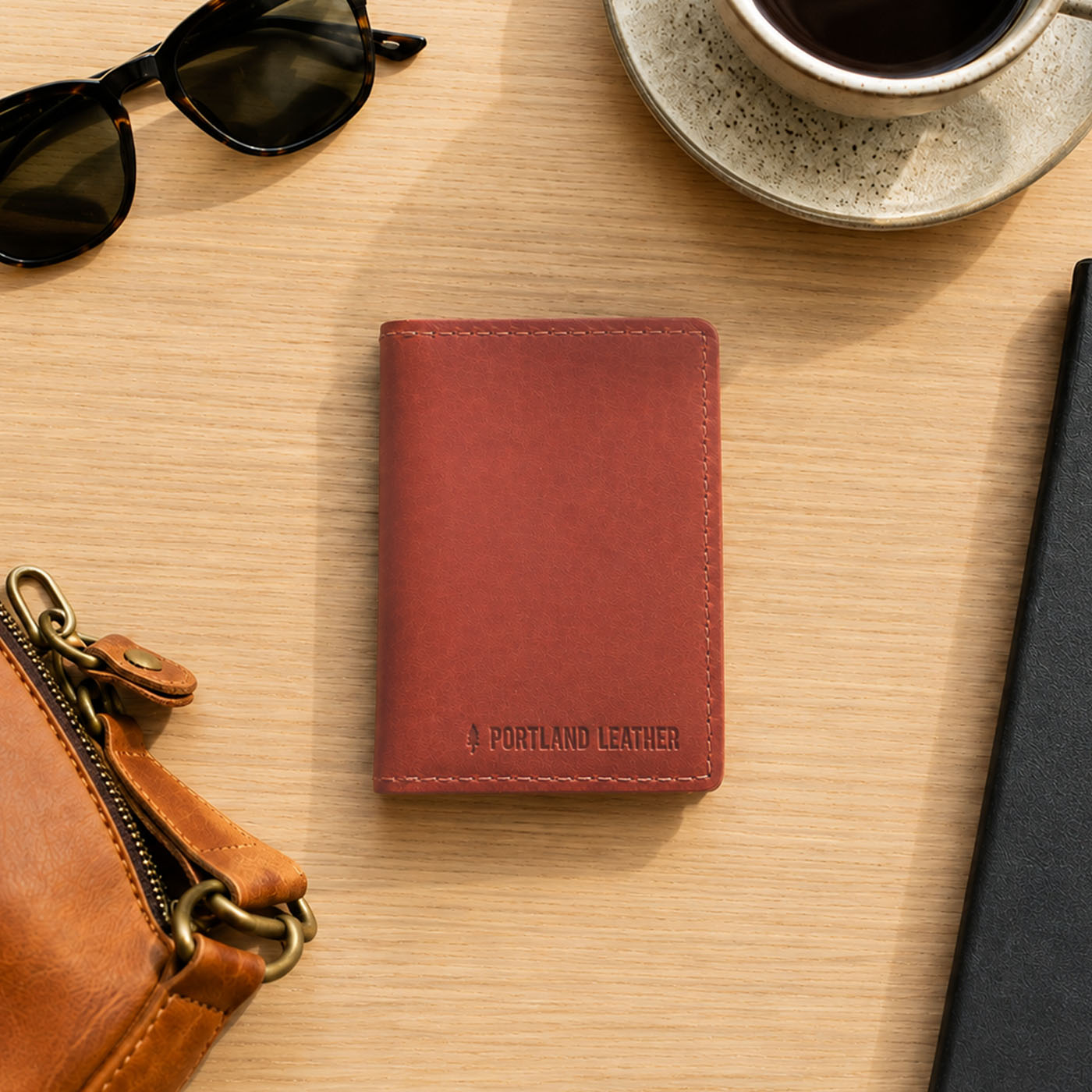 Marrone | A leather card holder displayed on a light wood desk with sunglasses, a coffee cup, and a brown leather pouch