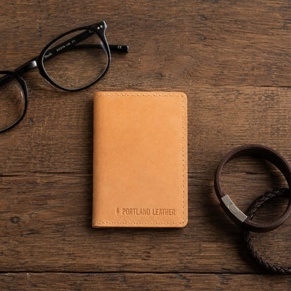 Naturale | A leather card holder styled on a rustic wooden tabletop with black eyeglasses and a dark leather bracelets