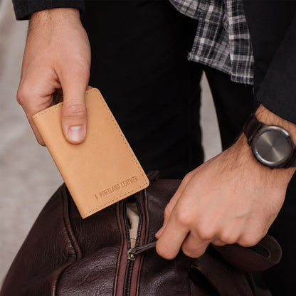 Naturale | A close-up lifestyle shot of a person's hands holding a card holder near a brown leather bag