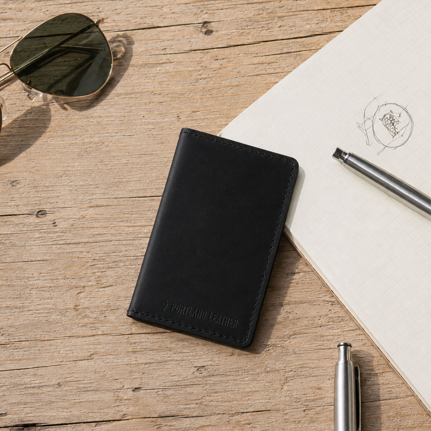 Nero | A leather card holder styled on a rustic wooden tabletop next to sunglasses, a notebook, and pens