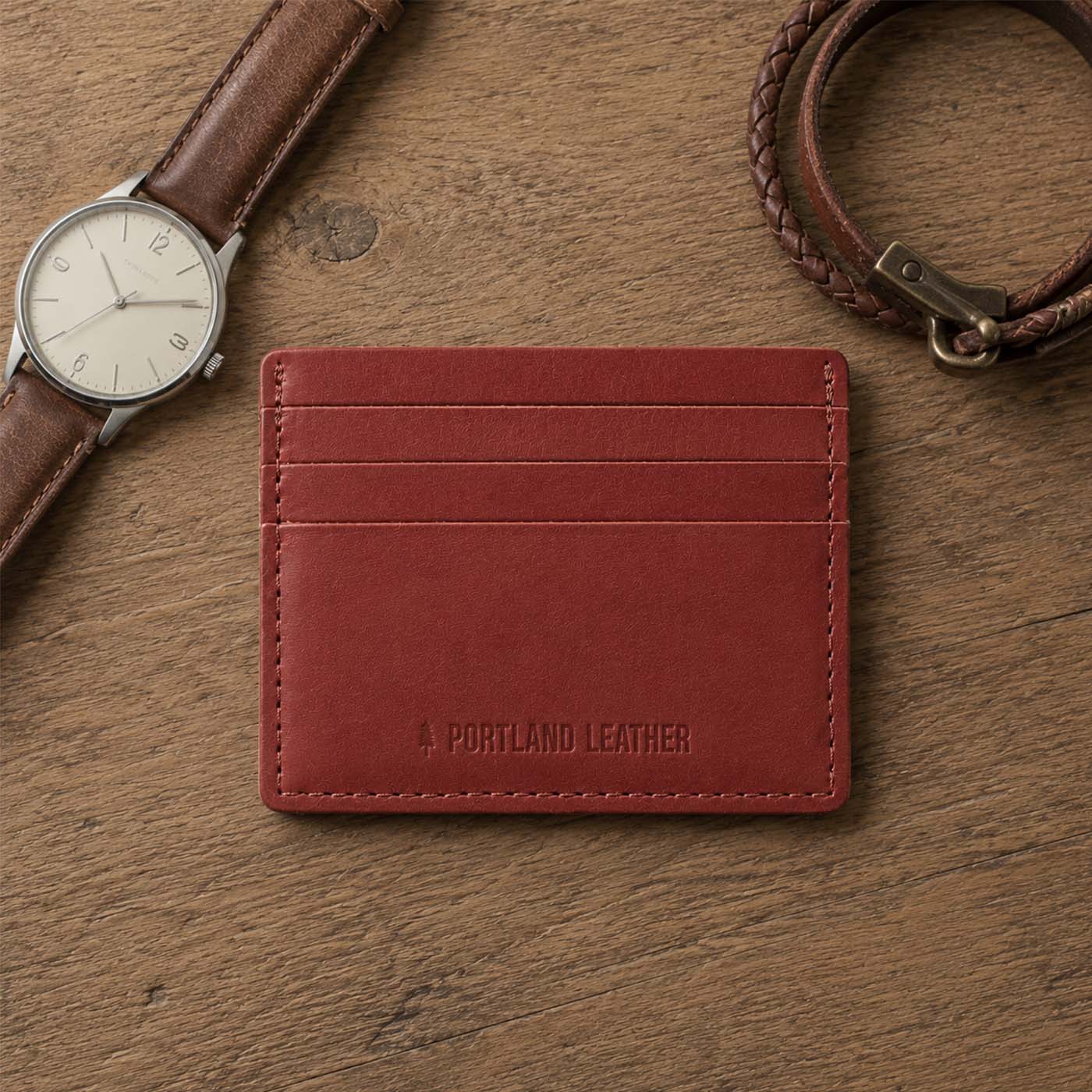 Marrone | A leather card holder displayed on a wooden surface next to a brown leather watch strap and a braided bracelet