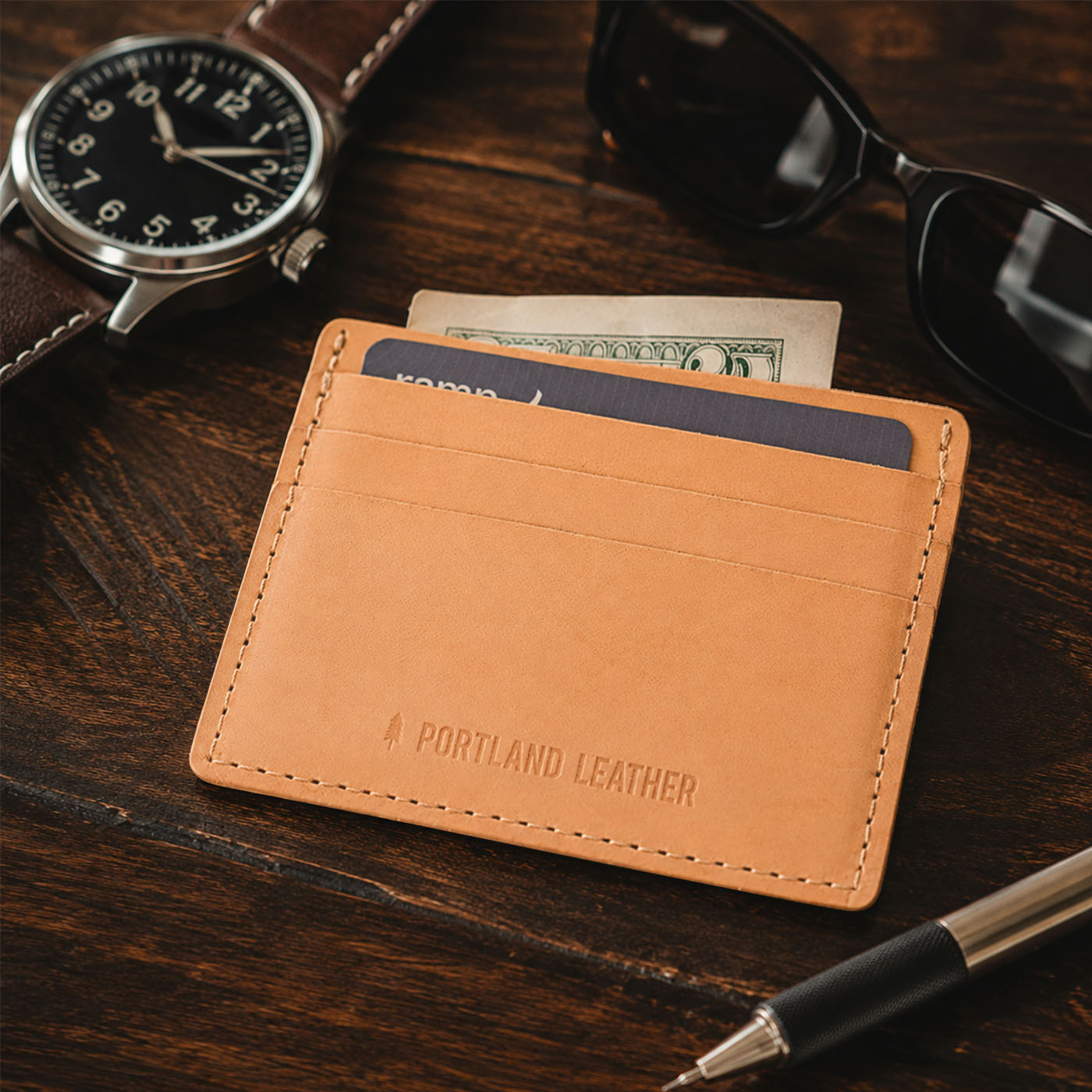 Naturale | A leather card holder shown on a dark wooden tabletop with a watch, sunglasses, cash, and a pen