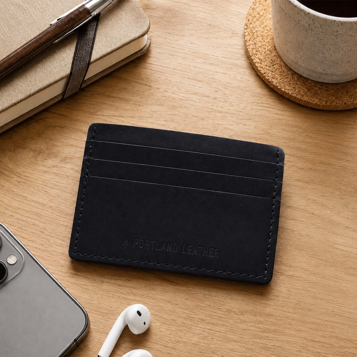 Nero | A leather card holder styled on a wooden desk with a notebook, pen, earbud case, phone, and coffee cup