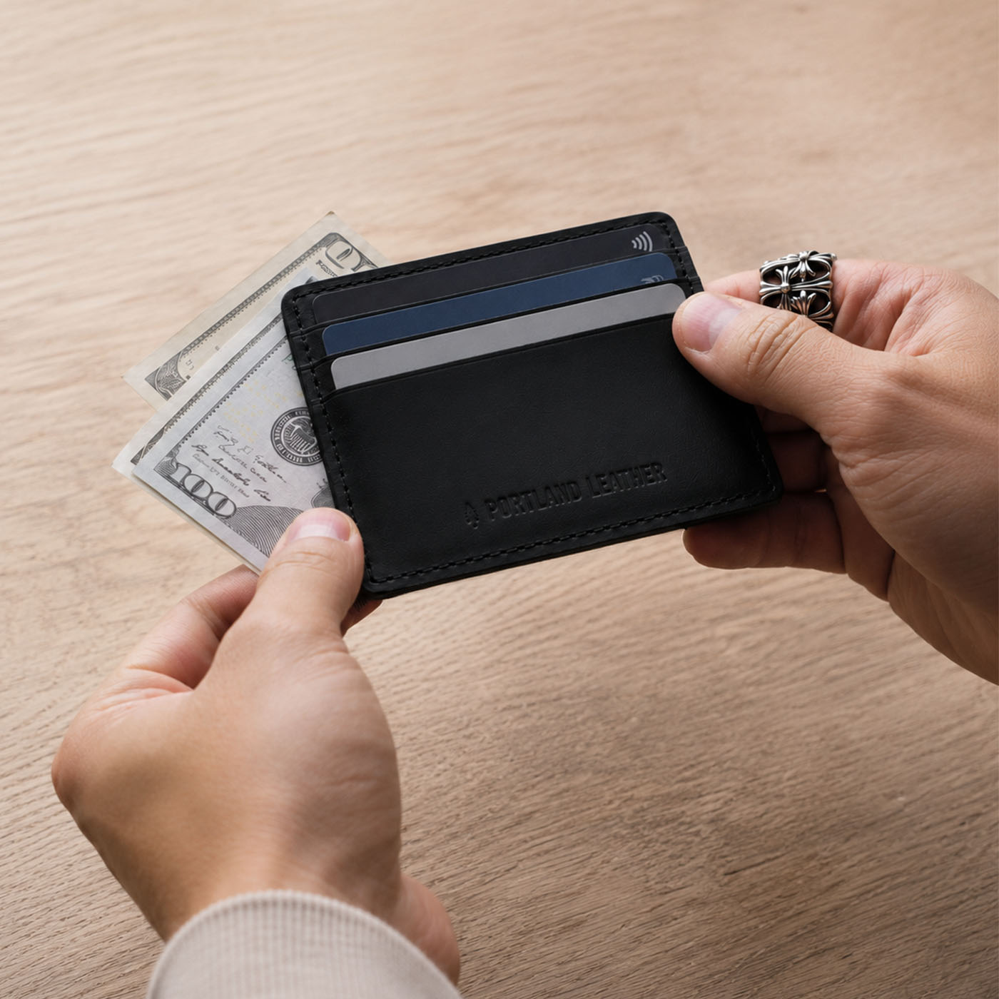 Nero | A card holder held by two hands over a light wood table, with two credit cards and a stack of cash partially visible inside