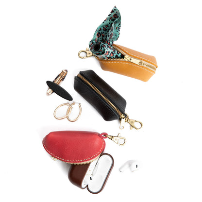 All Variants | Three taco shaped leather pouches with zippers and lobster clasps showing dog waste bags, hair clips, and AirPods inside